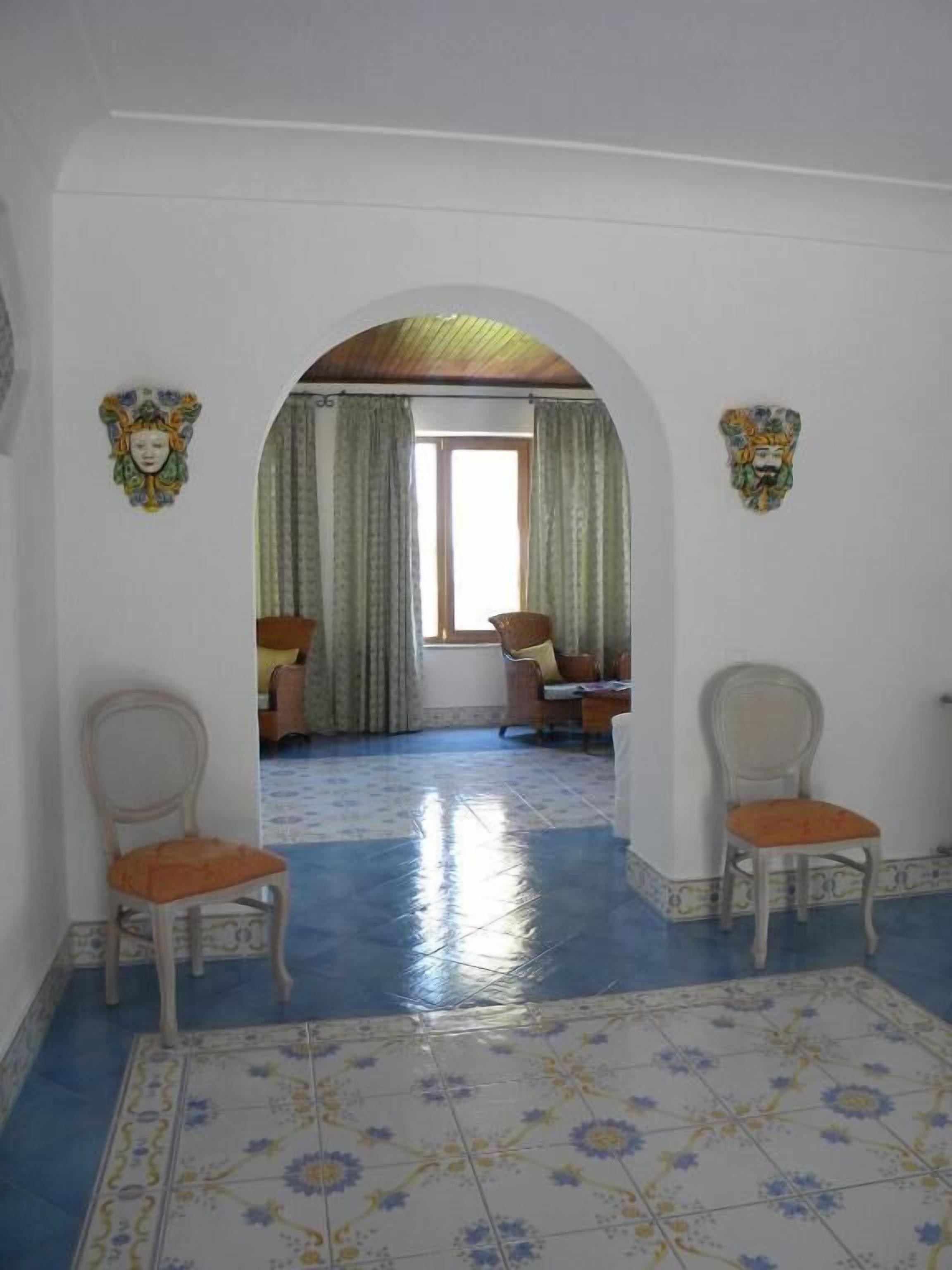 Une pièce avec un sol carrelé, deux chaises, une fenêtre avec des rideaux et des œuvres d’art décoratives accrochées au mur.