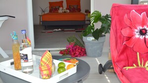 A hotel room with a bed, a table with a tray of fruits and drinks, a potted plant, and a red chair with a floral design. - Logement "paprika" 2 À 6 Pers (Le Gosier)