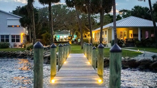 A wooden pathway leading to a building with a white exterior, surrounded by palm trees and a body of water.