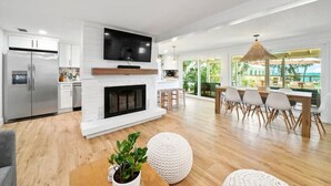 A modern living room with a fireplace, a dining area with a large table and chairs, and a kitchen with a refrigerator and cabinets.