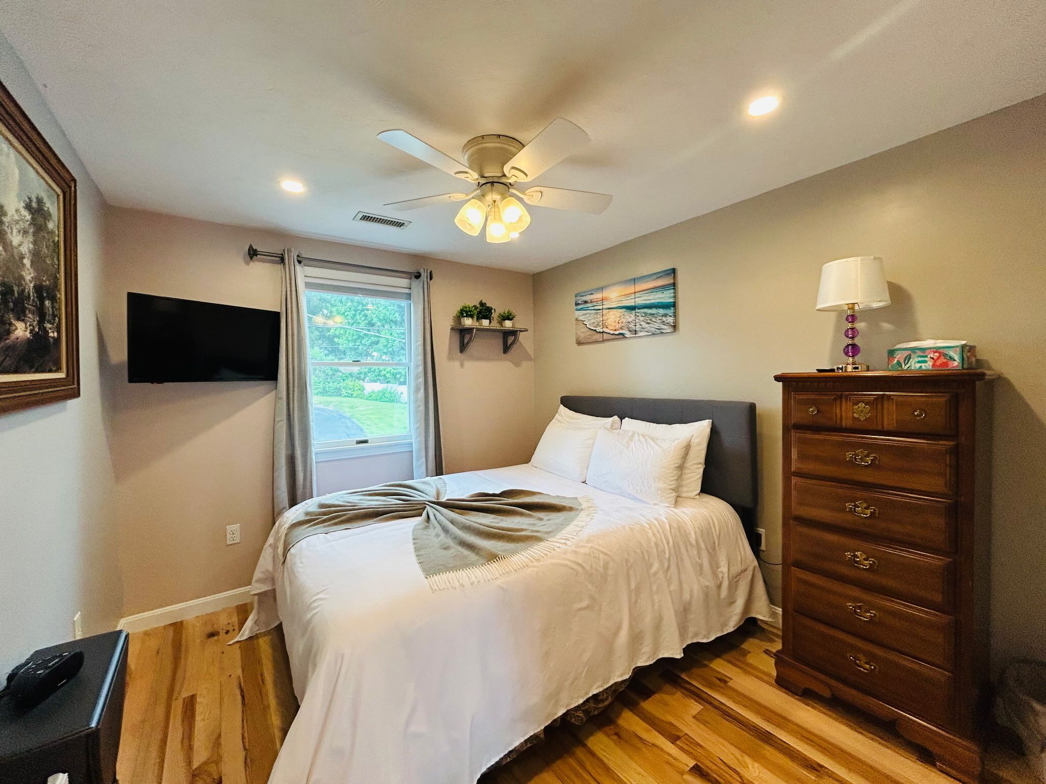 A bedroom with a bed, a ceiling fan, a dresser, a TV, a window, and a painting.