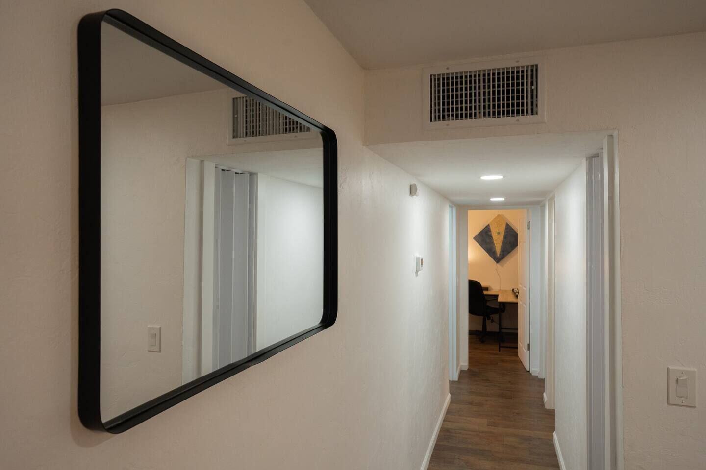 A hallway with a large mirror, a vent, and a dining area in the background.