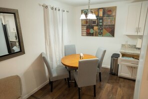 A dining area with a round wooden table, four grey chairs, a white curtain, a painting on the wall, and a kitchen area with cabinets and a countertop.
