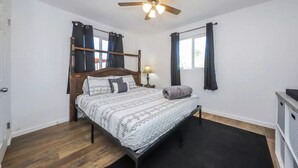 Une chambre à coucher avec un lit en bois, un ventilateur de plafond, une table de chevet avec une lampe et une fenêtre avec des rideaux.