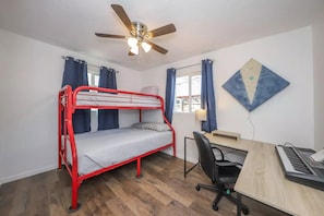 A room with a bunk bed, a desk with a computer, a chair, a ceiling fan, and a window with curtains.