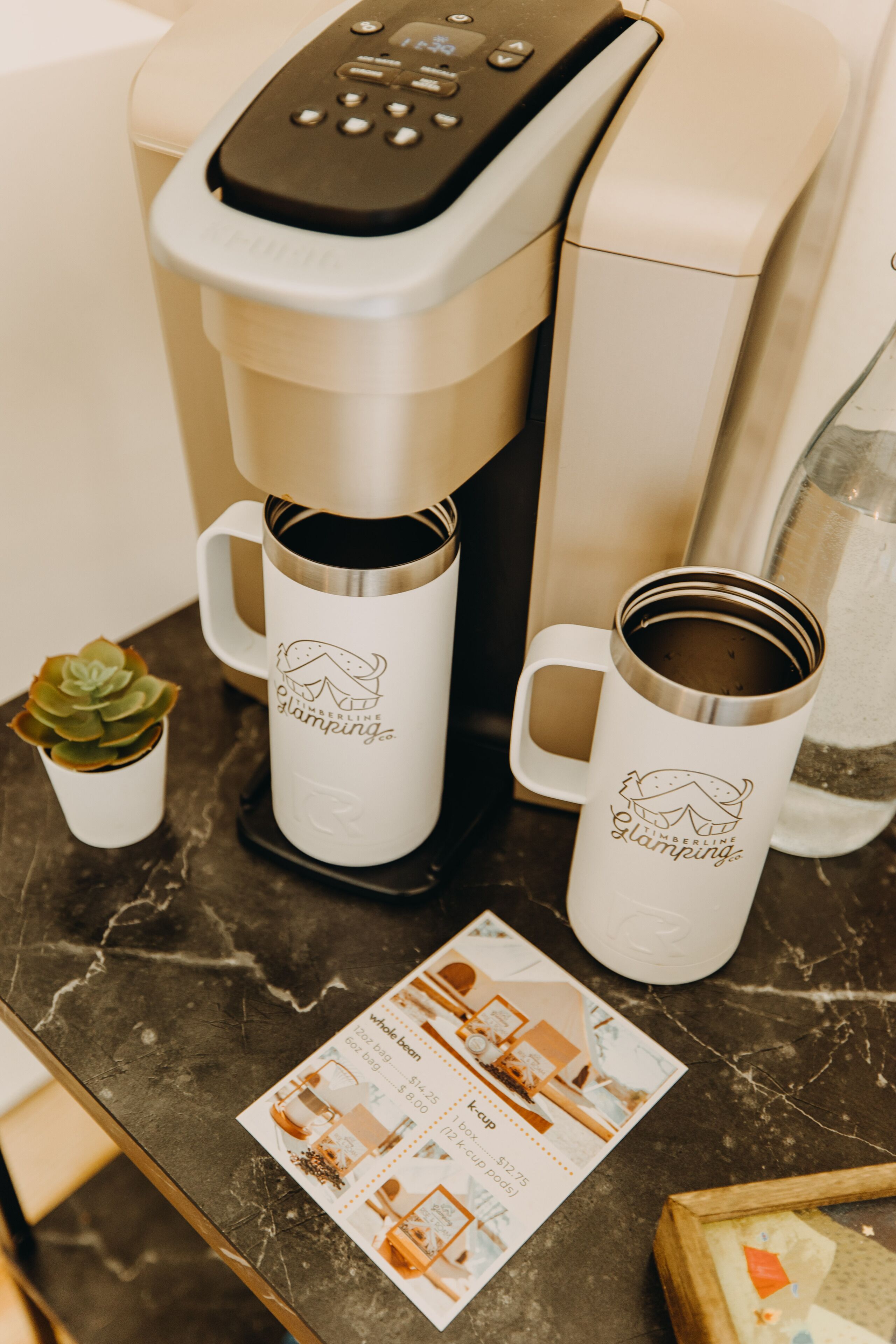A coffee maker, two mugs, a small plant, and a menu on a marble countertop.
