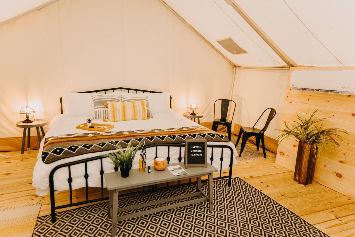 A tent-style bedroom with a bed, bedside tables, a chair, a small table with a sign, and a potted plant.