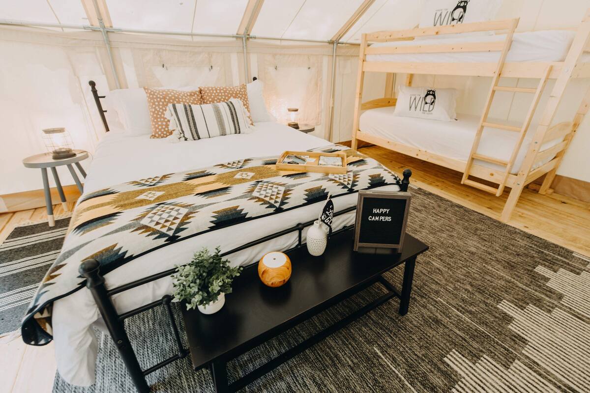 A tented accommodation room with a bed, a bunk bed, a bedside table with a tray, and a sign that reads "Happy Camper."