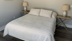 Una cama bien tendida con una colcha a rayas, dos mesitas de noche con lámparas, y un cuadro en la pared.