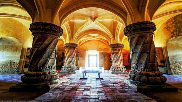 Interno di una cattedrale con soffitti a volta alti, grandi colonne in pietra e una navata centrale che conduce a una finestra luminosa.