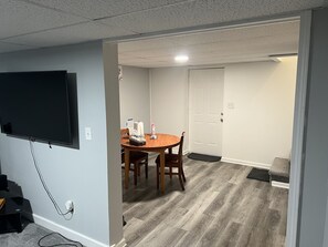 Una habitación con una mesa de comedor de madera, sillas, un televisor de pantalla plana montado en la pared y una puerta que da a otra habitación.
