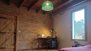 A room with a wooden floor, a desk with a chair, a bed with a red cover, a hanging green light fixture, and a window with a view of trees and a fence. - Maison Landaise Rénovée au Milieu de 2 Hectares (Sanguinet)