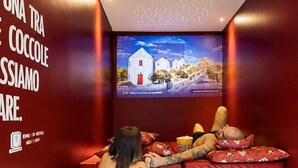 Uma mulher em uma banheira com uma tela de TV exibindo uma vista panorâmica, uma parede vermelha com uma mensagem e o logotipo de EMY IK HOTELS.