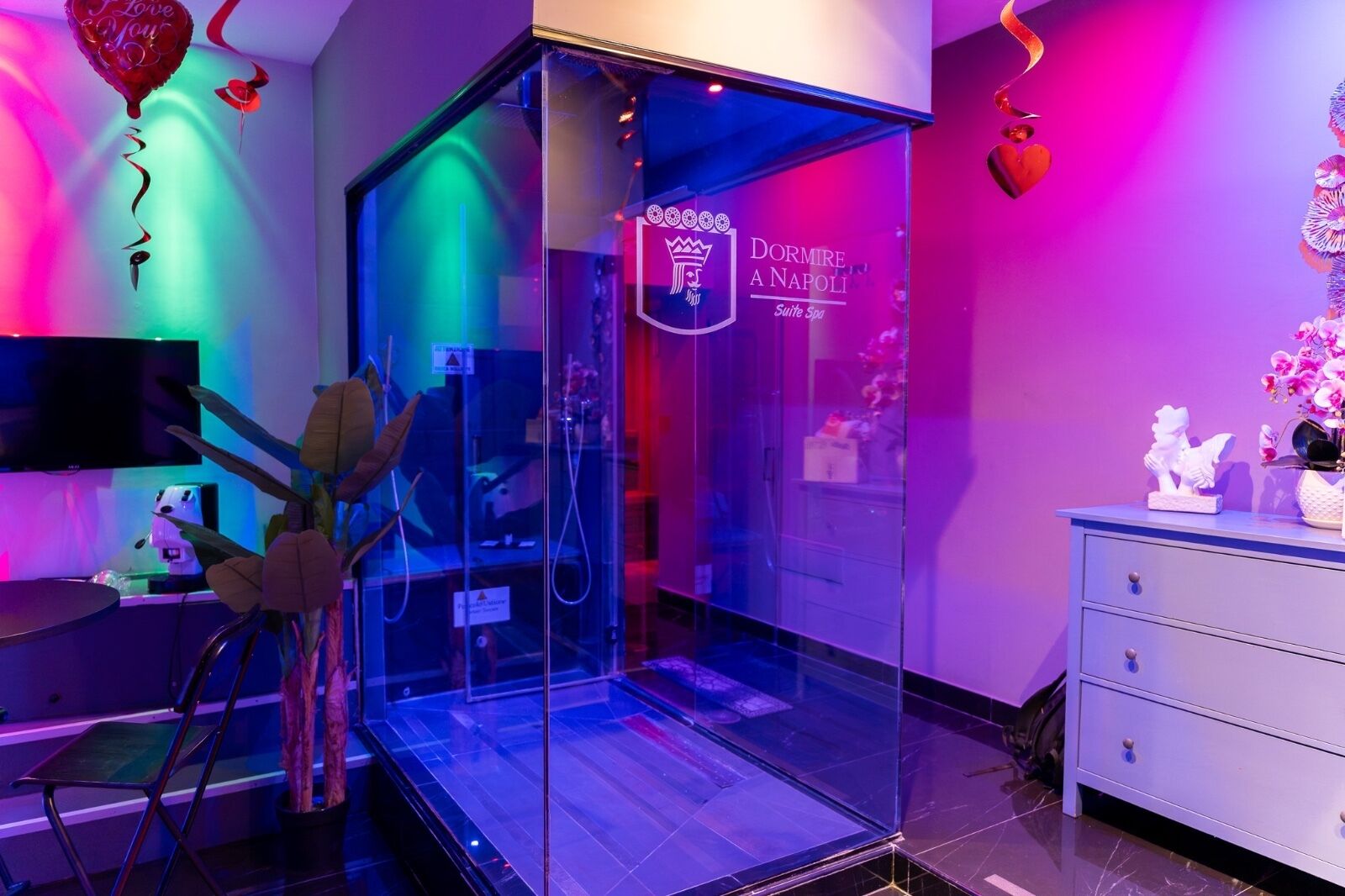 A hotel room with a glass partition, a sign reading 'Dormire a Napoli Suite Spa', a television, a desk with a chair, and decorative items including flowers and a heart-shaped balloon.