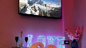 Quarto com televisão exibindo paisagem de montanhas, prateleira com garrafas e uma toalha em formato de coração, e um letreiro com a inscrição 'I LOVE'.