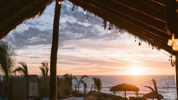 A beachside seating area with a thatched roof, overlooking the ocean at sunset. - Toninas México (Elota)