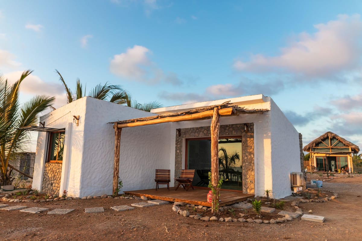 Un edificio blanco con una terraza de madera, paredes de piedra y una estructura de techo de paja al fondo.
