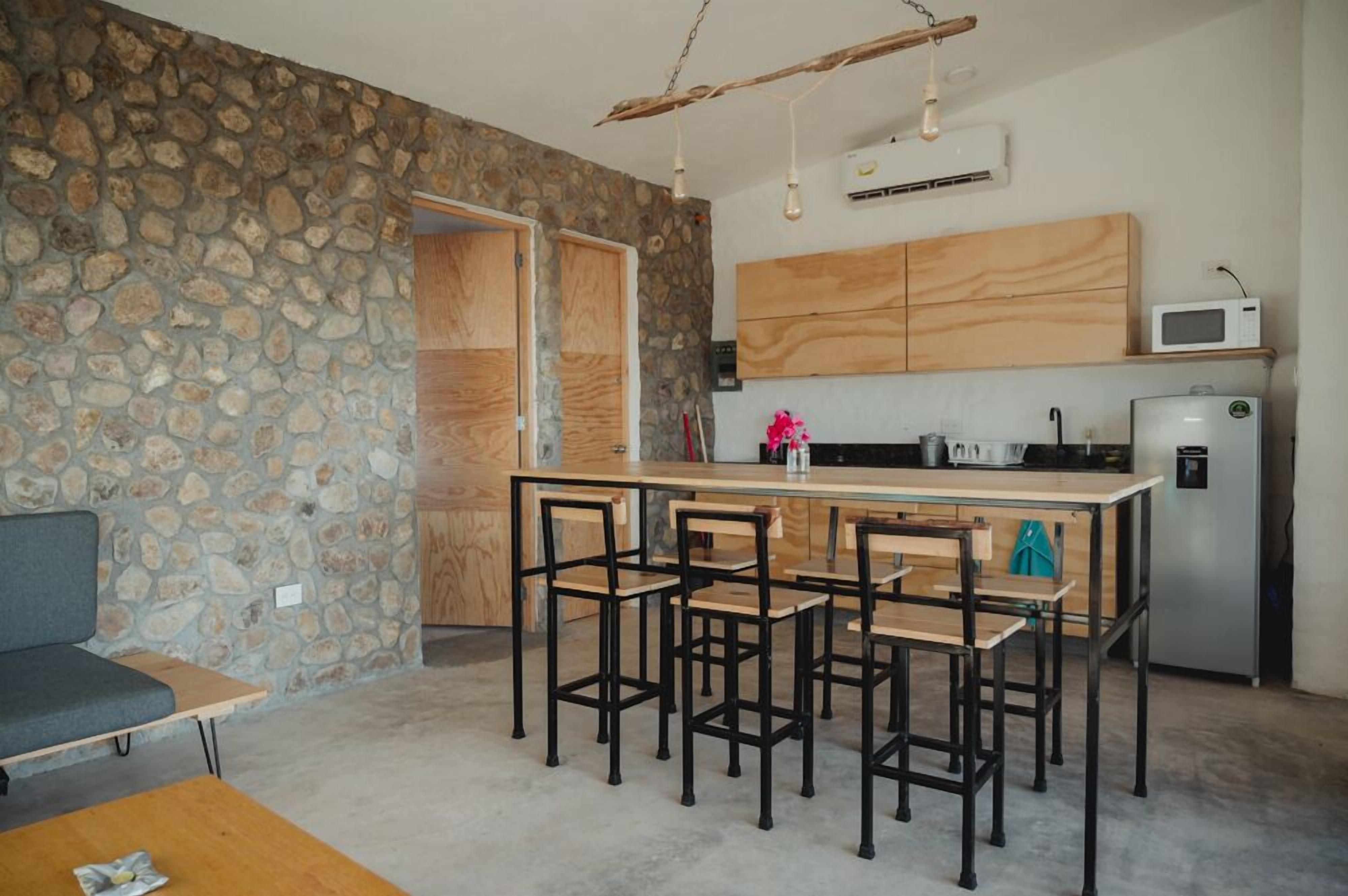 Una cuina compacta amb paret de pedra, armaris de fusta, una illa central i tamboretes de bar, i un microones.
