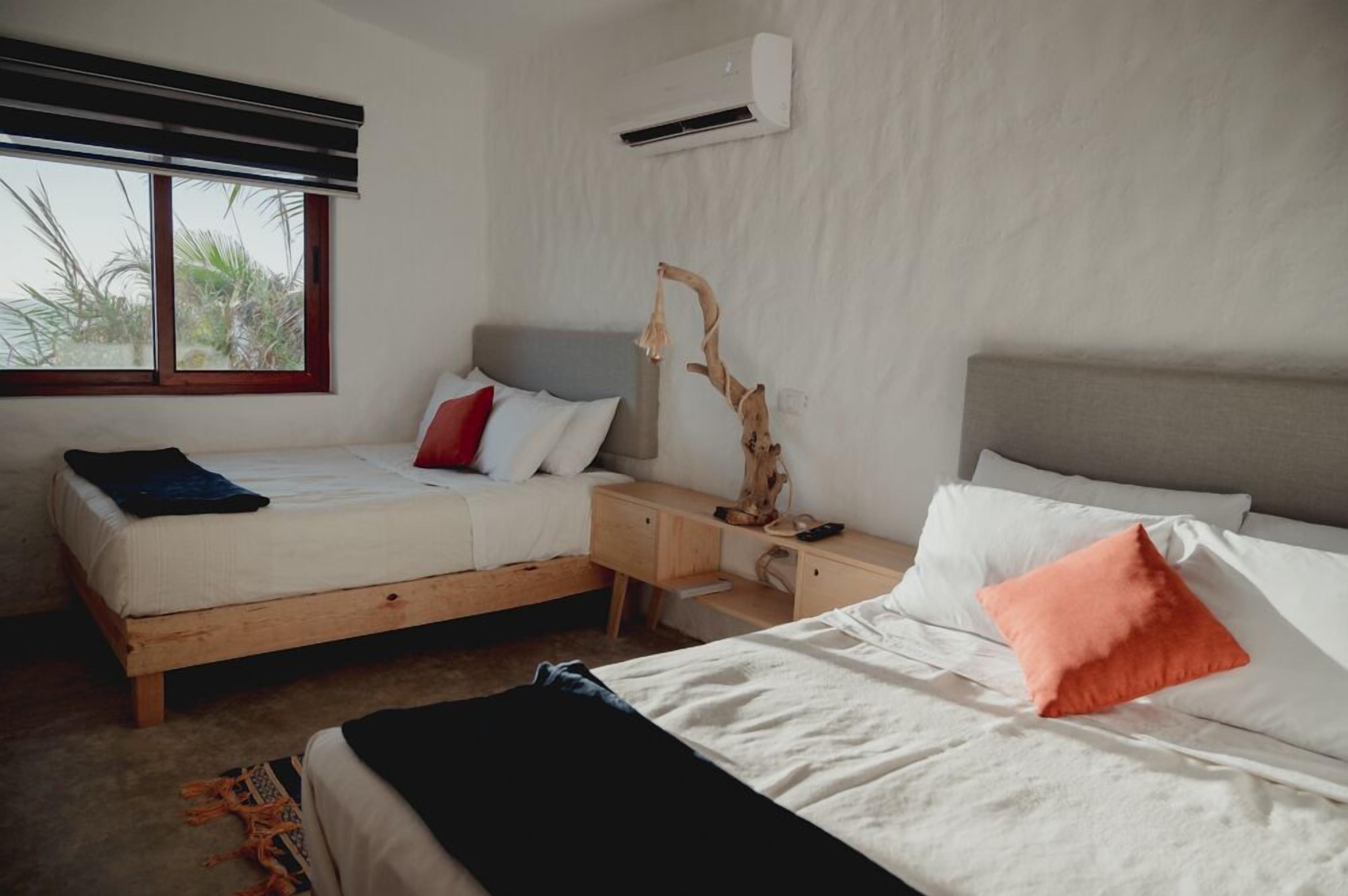 A bedroom with two beds, a wooden nightstand, an air conditioning unit, and a window with a view of palm trees.