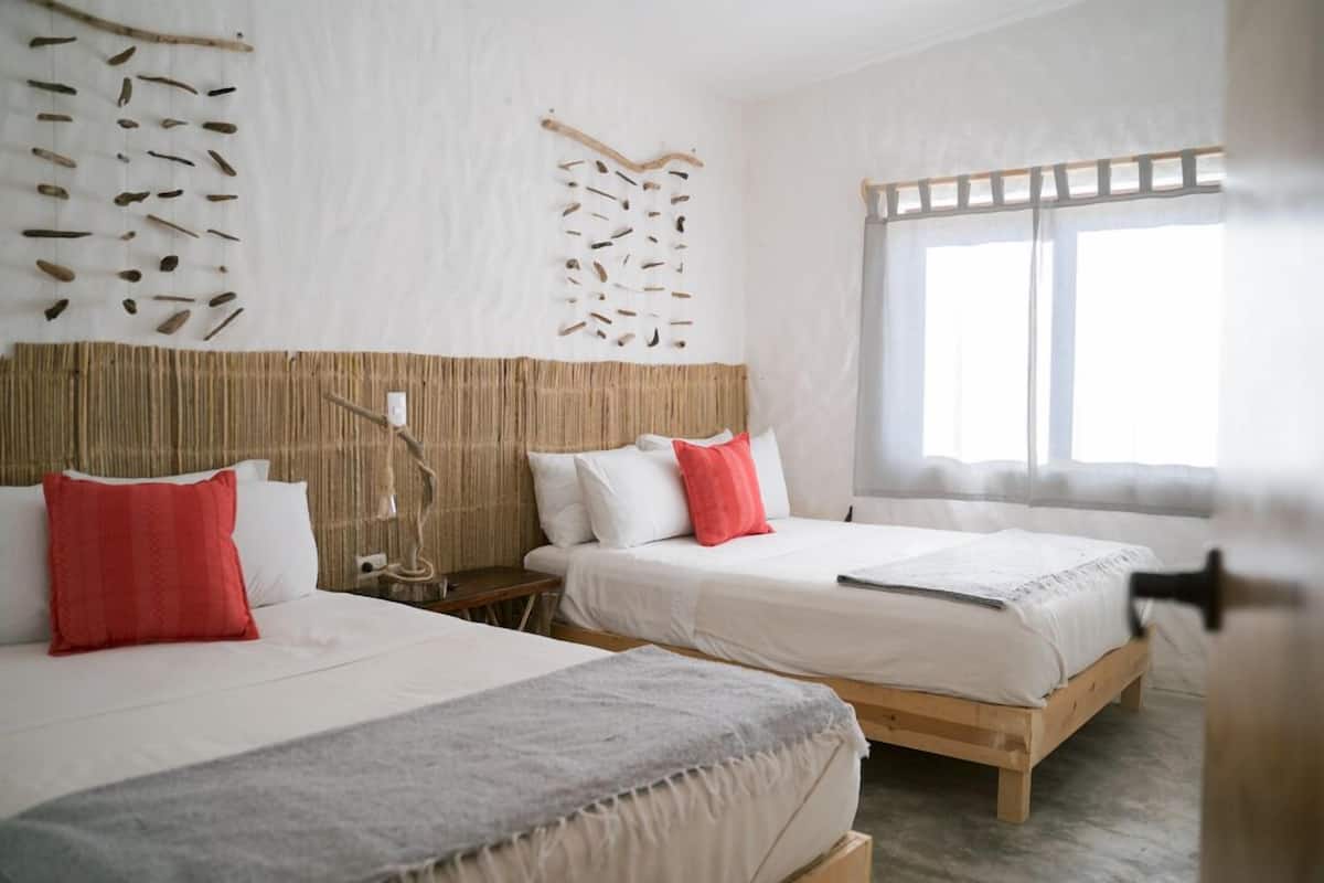 Habitación de hotel con dos camas, un ventanal grande y un muro decorado con ramas colgantes.