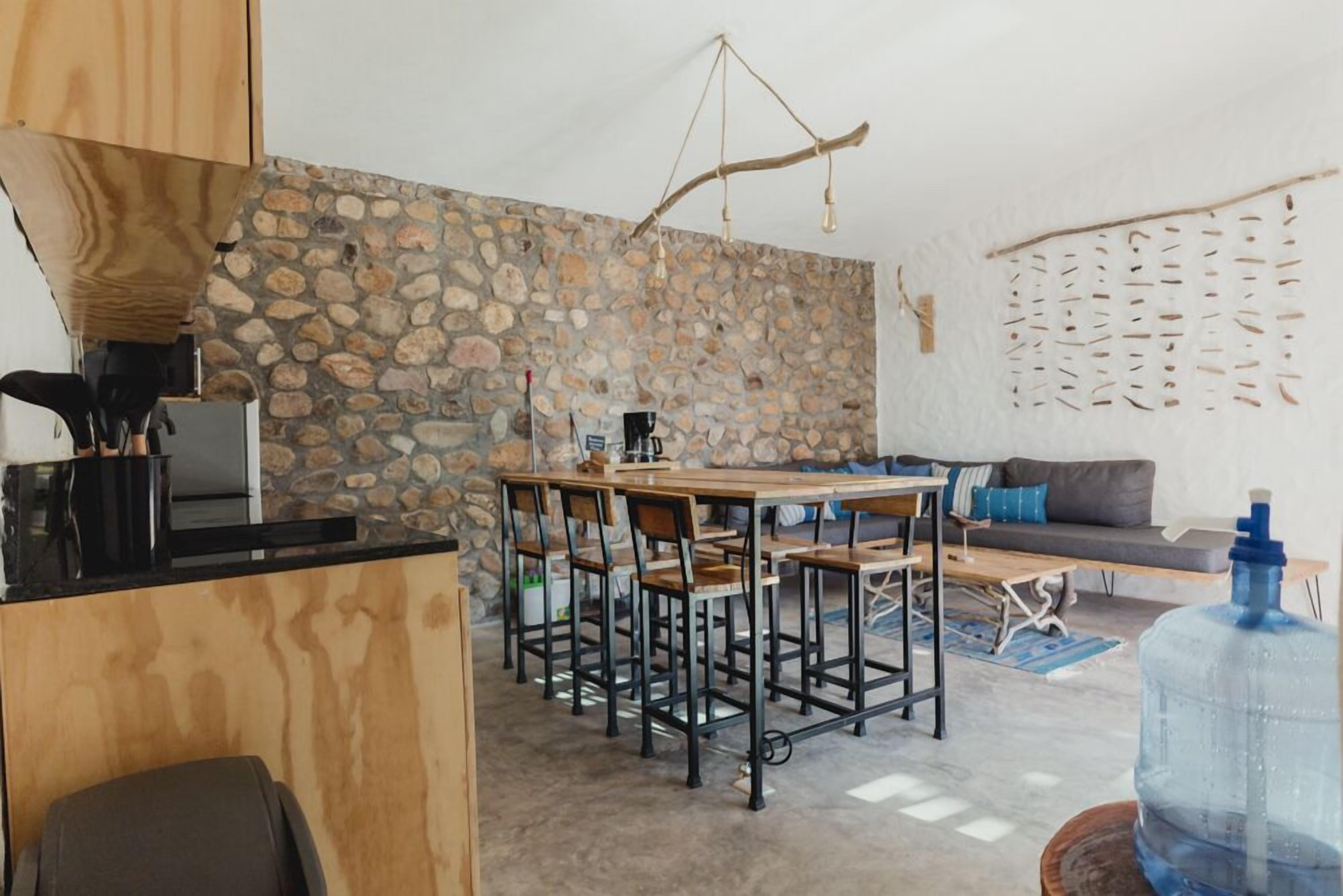 A modern kitchen with a stone wall, a wooden dining table with bar stools, a large water dispenser, and a sofa in the living area.