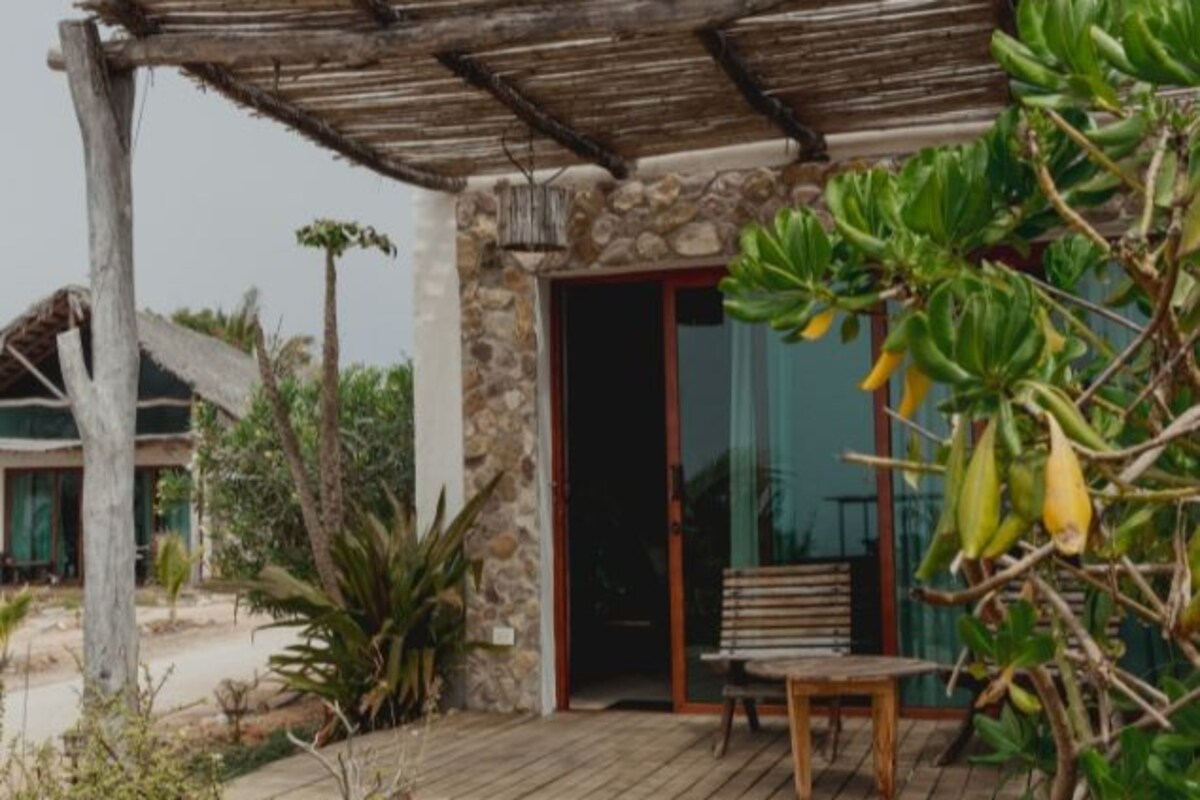 A rustic outdoor seating area with a wooden bench and a stone pathway leading to a building with a stone facade and a wooden pergola.