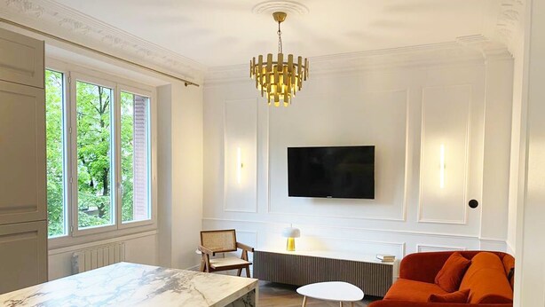Un salon moderne avec un comptoir en marbre, un canapé rouge, un téléviseur fixé au mur et un lustre.