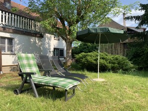 Uma área externa com gramado, com duas cadeiras de descanso, uma delas com almofada verde e branca xadrez, e um guarda-sol verde.
