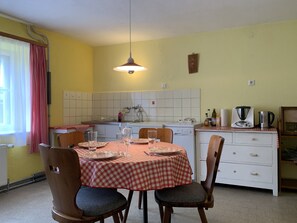 Uma área de refeições com toalha xadrez, cadeiras de madeira, luminária pendente, cozinha ao fundo e um rádio antigo.