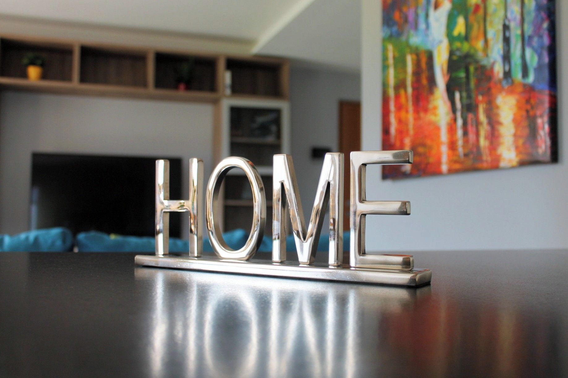 A metallic sign spelling "HOME" on a reflective surface, with a blurred background featuring a fireplace and a colorful abstract painting.