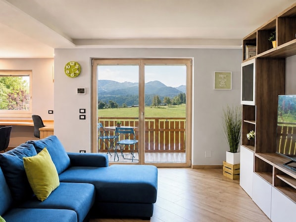 Un salon avec un canapé bleu, un meuble TV en bois, un coin repas avec une table et des chaises, et un balcon offrant une vue sur la montagne.