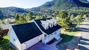 Une maison à deux étages, avec un toit sombre, une véranda et un garage. Elle est entourée d’une zone boisée et on aperçoit une montagne au loin.