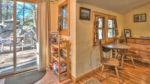 Ein gemütliches Zimmer mit einem Holztisch, Stühlen und einem Bücherregal. Große Fenster mit Vorhängen lassen natürliches Licht und Ausblicke ins Freie herein.