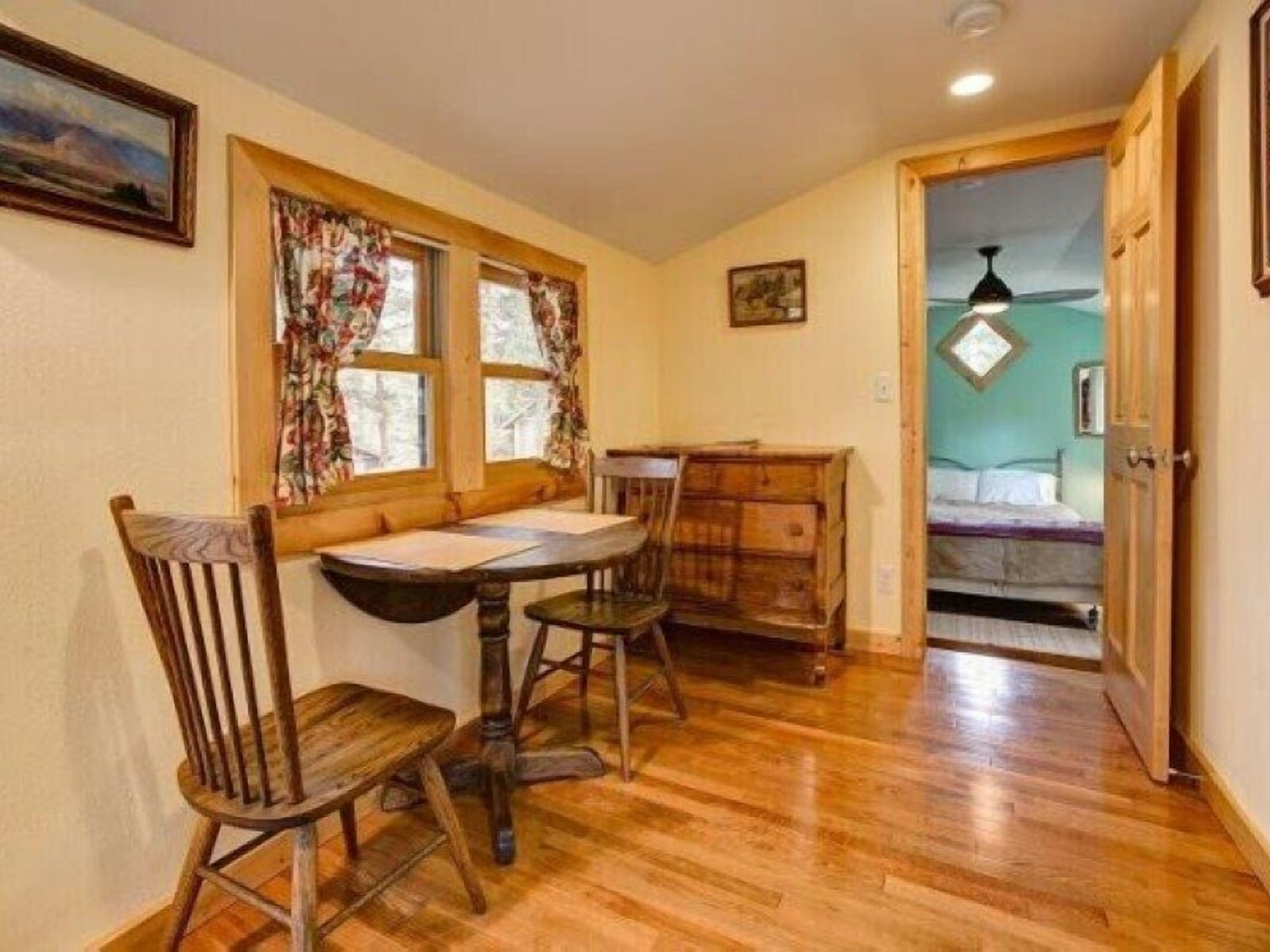 A cozy room with a wooden dining table, two chairs, a small wooden cabinet, and a bed in the adjacent room.
