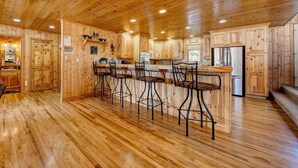 A wooden cabin interior with a kitchen area featuring wooden cabinets, a bar with stools, and a refrigerator.