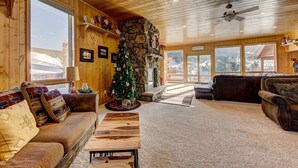 A cozy lodge interior with a stone fireplace, wooden furniture, and a large window overlooking a snowy landscape.