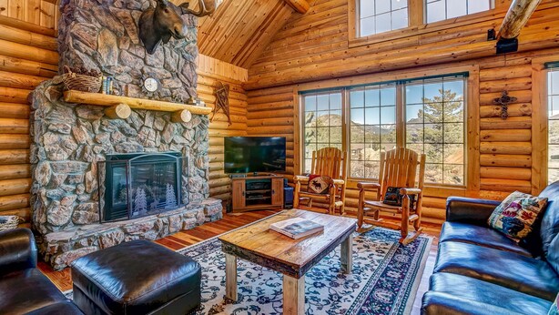 A log cabin interior with a stone fireplace, wooden furniture, and large windows offering a view of the outdoors. - Moose Mountain Lodge - Secluded Log Home w/Beautiful Mountain Views (Estes Park)