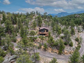 Une cabane dotée d’une terrasse en bois, entourée d’une forêt dense et d’un terrain rocheux, sous un ciel dégagé.