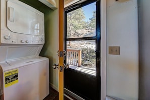 A laundry room with a washer and dryer, a window with a view of trees and rocks, and a door with a wooden railing. - Ridge View - Mountain Top Home with 360 Views (Estes Park)