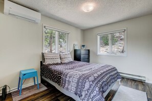 A bedroom with a bed, a dresser, a small blue table, and two windows with a view of trees.