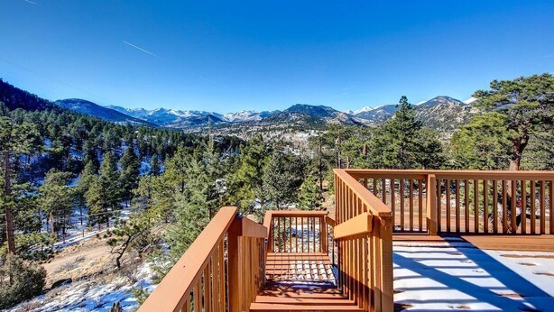 A wooden staircase leading to a deck with a view of snow-capped mountains and a forested valley.