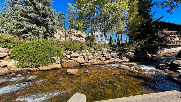 A clear stream with rocks and trees, a stone bench, and a building in the background. - Stone's Throw - A Downtown Estes Park getaway (Estes Park)