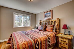 Une chambre avec une grande fenêtre, une tête de lit en bois, une couvre-lit à motifs, une table de chevet avec une lampe et un tableau encadré au mur.