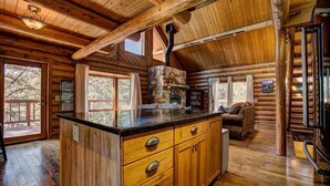 A log cabin interior with a stone fireplace, wooden cabinets, and a large window overlooking a forest. - Windham Drive - Beautiful Secluded Mountain Home (Estes Park)