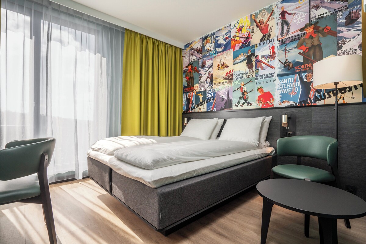 A modern hotel room with a bed, a desk, a chair, and a large wall mural.