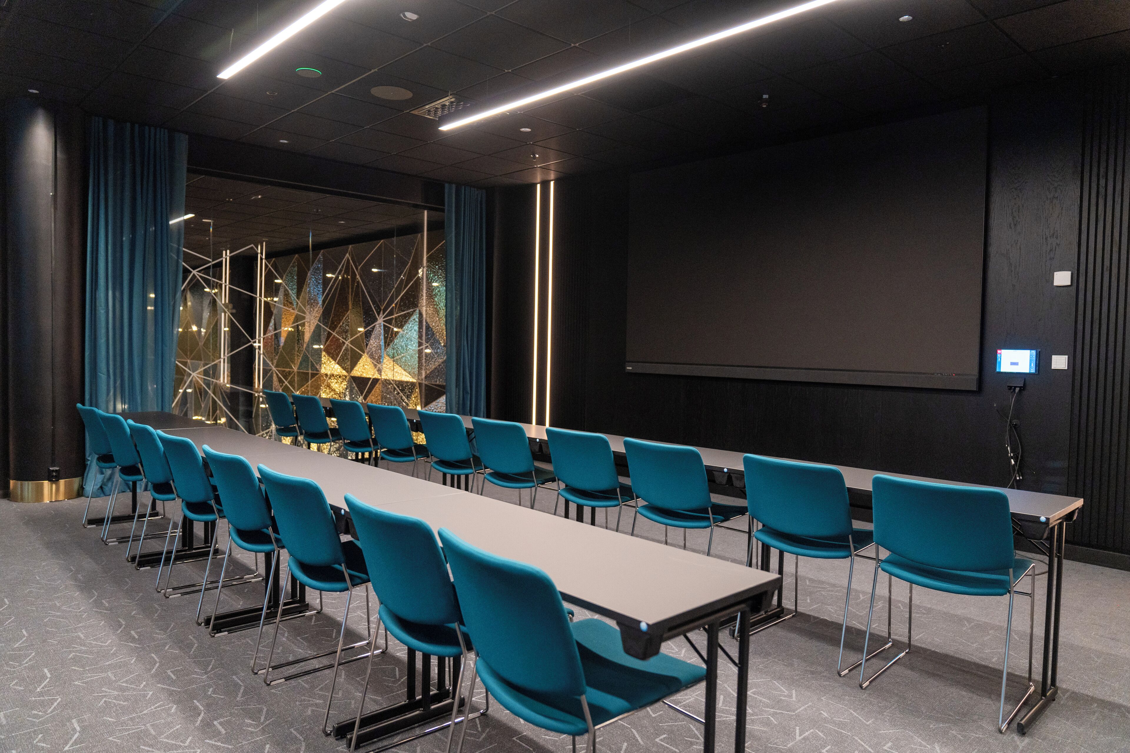 Una sala de conferencias con una larga mesa, sillas azules, una pantalla de proyector y un ventanal con un diseño geométrico.