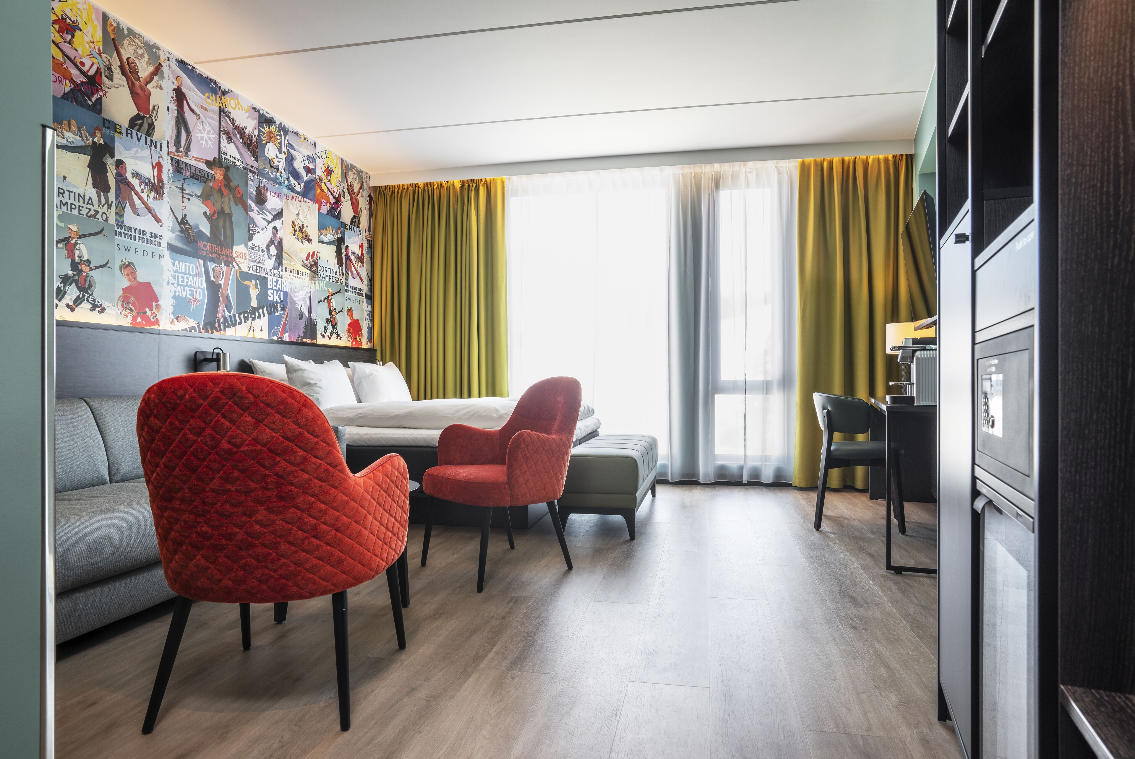 A modern hotel room with a bed, a red chair, a grey sofa, and a dining area.