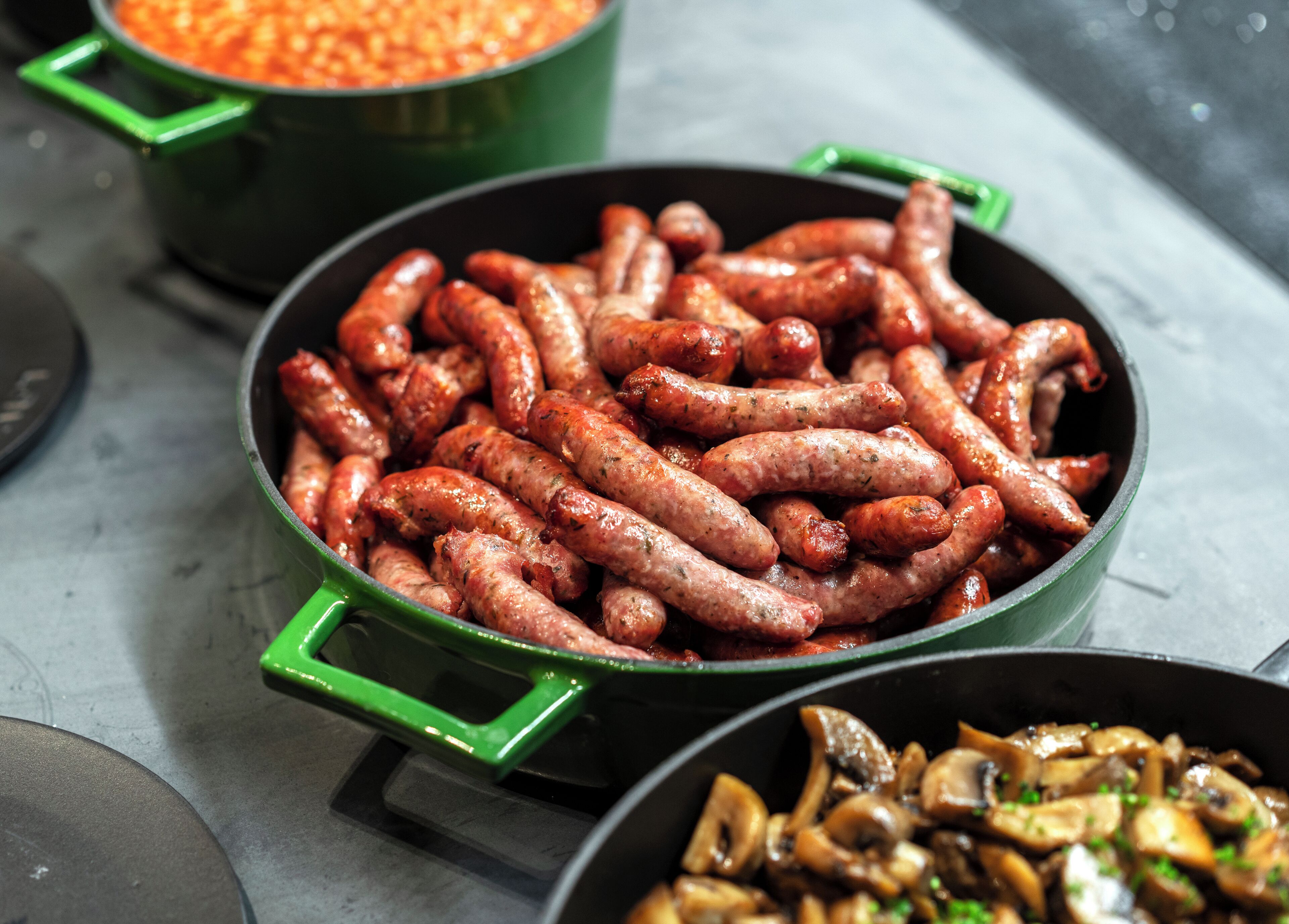 A close-up of cooked sausages in a green pan, with a bowl of mushrooms in the foreground.