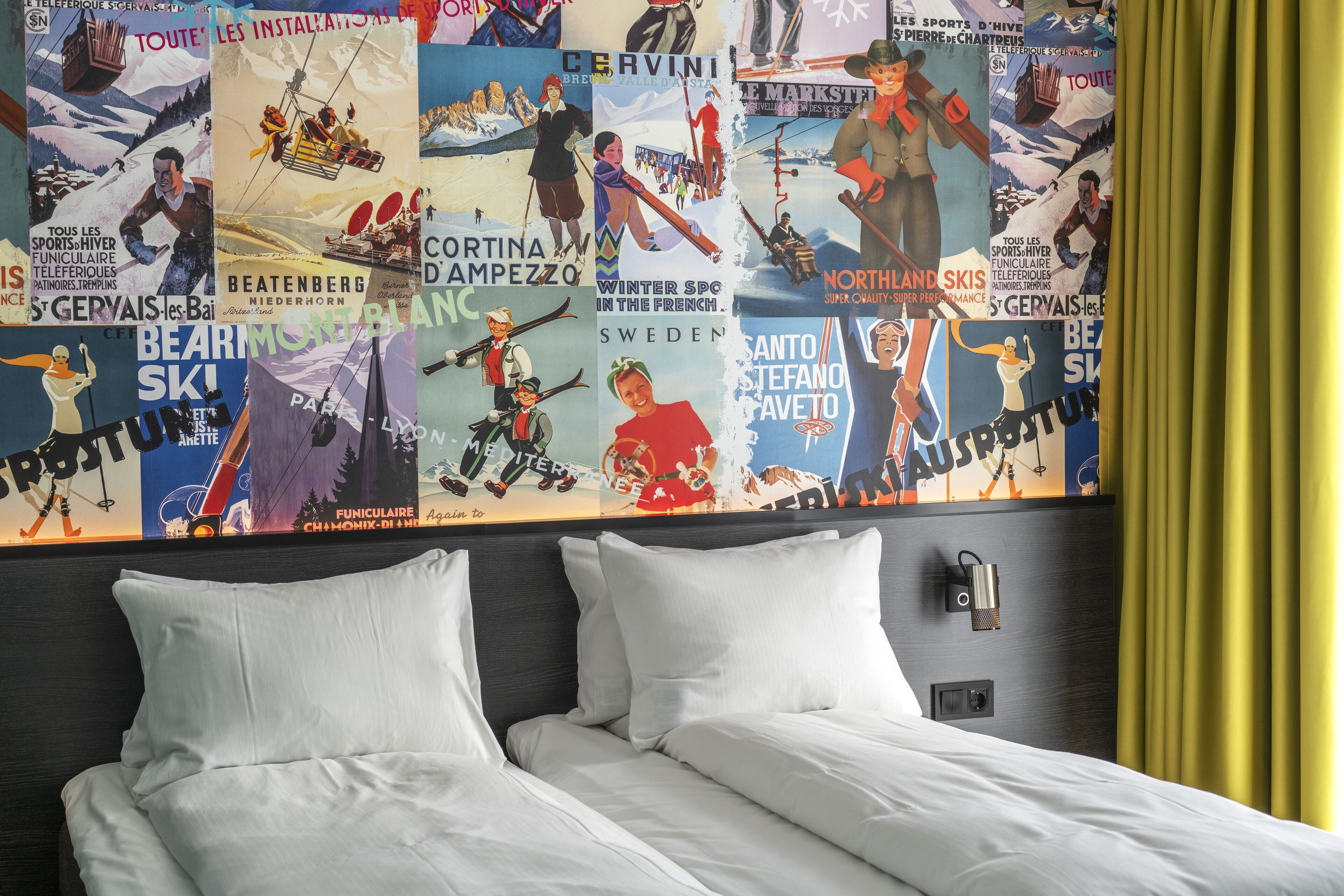 A hotel room with a bed, white pillows, and a wall decorated with ski-themed posters.