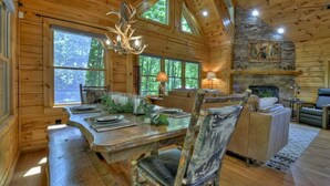 A rustic wooden cabin interior with a dining table set for four, a fireplace, and large windows offering a view of the forest. - Cedar Ridge | Hot Tub + Game Room + Fire Pit (Blue Ridge)